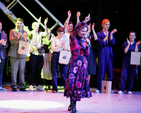 Na zdjęciu Joanna Szczepkowska stojąca na scenie. Ubrana w długą, ciemną sukienkę w czerwone kwiaty. Idzie podnosząc dłoń do góry w geście wiktorii, uśmiecha się. W tle w rzędzie stoją kobiety i mężczyźni, biją brawo. Niektórzy z nich trzymają w dłoniach jednakowe papierowe torby z ciemnym logotypem z literą T. Światła na scenie są kolorowe.